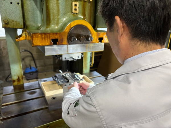 工場内で作業服を着た男性が、大型の産業機械（プレス機と見られる）を操作している様子を後ろから捉えた画像です。機械の作業台には金属部品や木箱が置かれています。