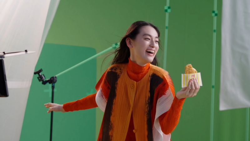 グリーンバックのスタジオで、オレンジ色の服を着た笑顔の女性がフライドチキンを手に持っています。