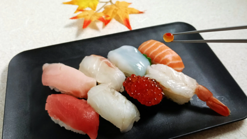 黒い皿に盛り付けられた色とりどりの握り寿司のセット。サーモン、マグロ、エビ、イカ、いくらなど様々なネタがあり、箸でいくらが一つ摘まれている。背景には紅葉の飾りが添えられている。