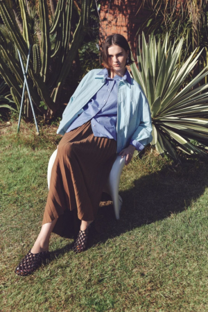 屋外の庭で白い椅子に座る女性の全身像です。水色のジャケットと青いストライプシャツ、茶色のプリーツスカートを着用し、背景にはサボテンなどの植物が写っています。自然光が差し込む中で、落ち着いた表情をしています。