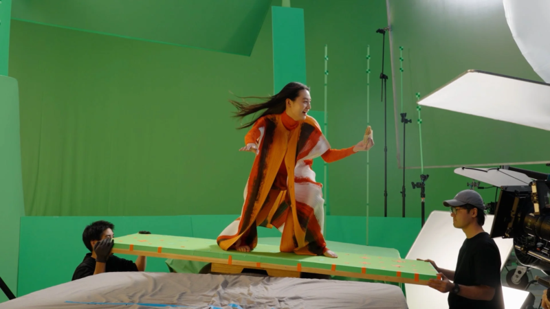 グリーンバックのスタジオで、カラフルな衣装を着た人物が傾斜した足場の上に立っています。