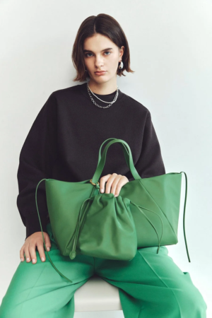 黒いトップスと鮮やかな緑のパンツを着用した女性が、同色の大きなトートバッグと小さなポーチを手にしています。シンプルな背景で、ショートヘアのモデルがアクセサリーを身につけ、カメラを見つめています。