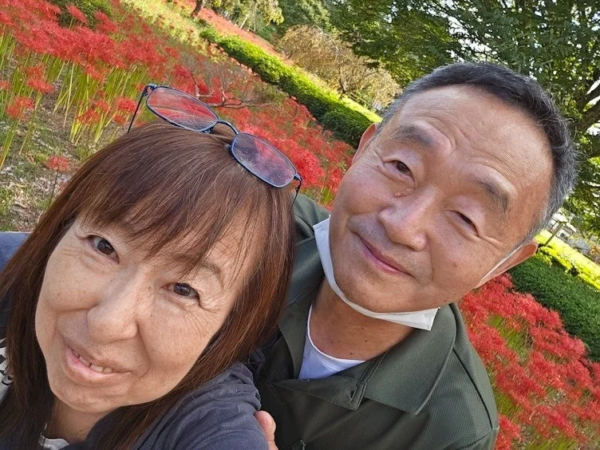 彼岸花が咲き誇る美しい背景の前で、笑顔のシニアカップルが楽しそうにセルフィーを撮っています