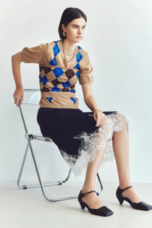 白い背景のスタジオで、透明な椅子に座る若い女性モデル。アーガイル柄の半袖ニットとネイビーのレーストリムスカートを着用し、アクセサリーを身につけている。落ち着いた雰囲気のファッションポートレート。
