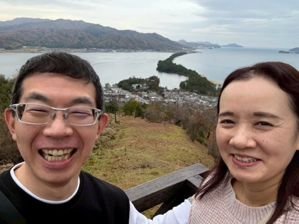 天橋立の美しい景色を背景に、笑顔でセルフィーを撮る男女二人の姿が写っています