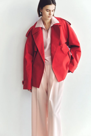 赤いジャケットとピンクのポロシャツ、淡いピンクのワイドパンツを着用した女性が白い壁の前でポーズをとっています。シンプルながらも色使いが特徴的なファッションスタイルです。