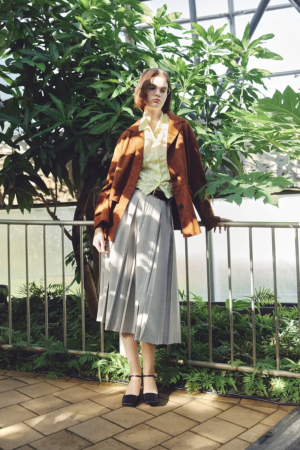 温室のような場所で、茶色のジャケットとグレーのプリーツスカートを着用した女性が立っているポートレート。緑豊かな植物が背景に広がり、自然光が差し込む中で、モデルが落ち着いた表情を見せている。