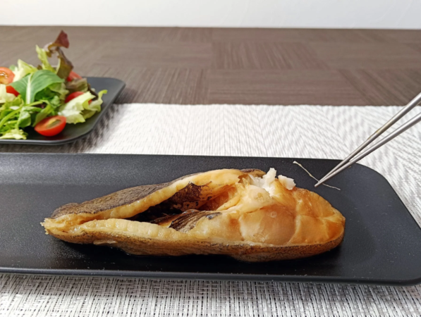 黒い皿に盛られた魚料理と、奥に添えられたフレッシュなグリーンサラダが写っています。箸が魚に触れており、美味しそうな和食の食卓風景です。