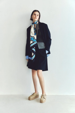 白壁を背景に立つ女性の全身ポートレート。ネイビーのブレザーとスカートにストライプシャツを合わせ、首元には青と白の柄スカーフを巻いています。ベージュのエスパドリーユを履き、洗練されたビジネスカジュアルな装いです。