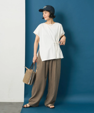 白い変形Tシャツとブラウンのワイドパンツを着用した女性が、ネイビーのキャップと麦わら素材のバッグを合わせたカジュアルな夏のコーディネートで立っています。
