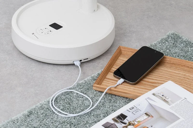 白い家電製品のベースからスマートフォンが充電されている様子。木製のトレイに置かれたスマホと雑誌が、緑色のカーペットとグレーの床の上に配され、現代的なライフスタイルの一場面を表現しています。