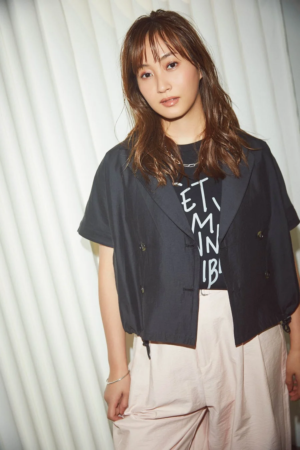 白い背景の前に立つ、ブラウンのロングヘアの若い女性のポートレート。彼女は黒い半袖ジャケットの下にグラフィックTシャツを着用し、淡いピンクのパンツを合わせています。