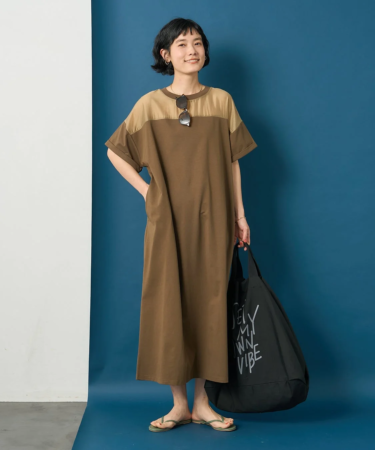 短い髪の女性が、ブラウンとベージュのツートンカラーのゆったりとしたマキシワンピースを着て、笑顔で立っています。