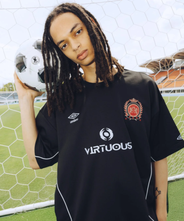 ドレッドヘアの男性が黒いサッカーユニフォームを着て、サッカーボールを抱え、サッカー場のゴールネットの前でポーズをとっています