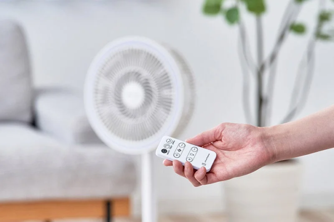 明るいリビングで、白い扇風機のリモコンを手に持っている様子です。快適な室内環境を操作するシーンを捉えています。
