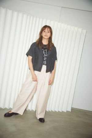 白いプリーツ状の壁を背景に、笑顔で立つ若い女性の全身ポートレート。黒いショート丈のトップスとTシャツ、ピンクベージュのワイドパンツを合わせた、カジュアルながらも洗練されたファッションが特徴です。