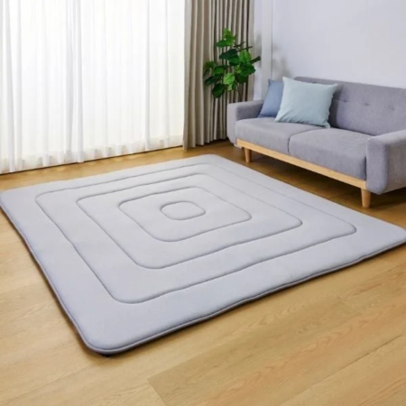 明るいリビングルームの床に、グレーのキルティング加工された四角いデザインのラグが敷かれ、奥には同系色のソファと観葉植物が配置されたモダンなインテリアの写真