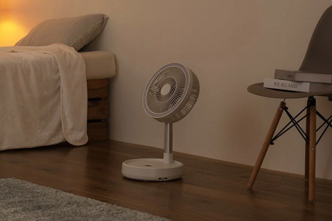 暖色系の照明が灯る部屋に、折りたたみ式の扇風機が置かれています。ベッドと椅子があり、シンプルでモダンなインテリアが落ち着いた雰囲気を醸し出しています。