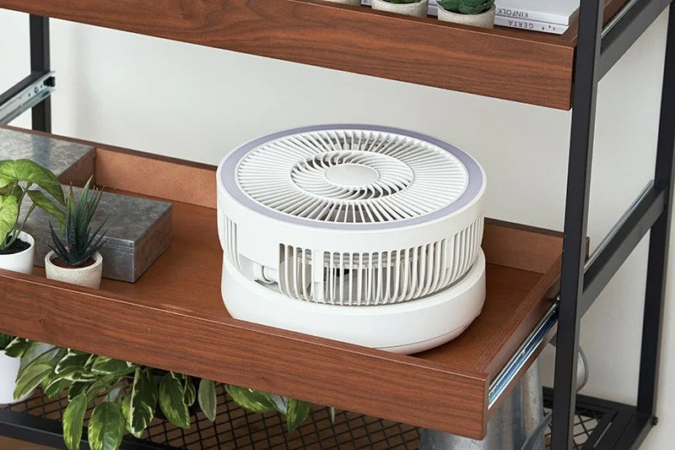 木製の棚に置かれた白いコンパクトな卓上扇風機が写っています。周囲には小さな鉢植えの植物が配置されており、モダンなインテリアの一部として機能しています。