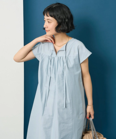 淡い水色のワンピースを着たショートヘアの女性が、青と白の背景の前でポーズをとっています。