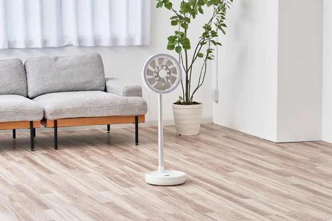 リビングルームに置かれた白いモダンな扇風機と、グレーのソファ、観葉植物が写っています。明るい木目のフローリングと白い壁が特徴の、すっきりとした空間です。