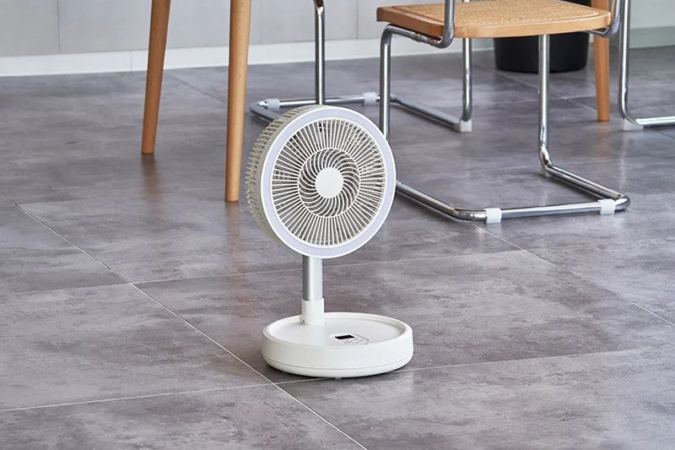 グレーのタイル張りの床に、LEDライトリング付きの白いポータブル扇風機が置かれています。背景には木製のテーブルと金属製の椅子の脚が見えます。