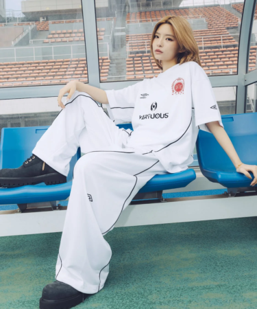 白いスポーツウェアを着た女性が、スタジアムの観客席にある青いベンチに座ってポーズをとっています