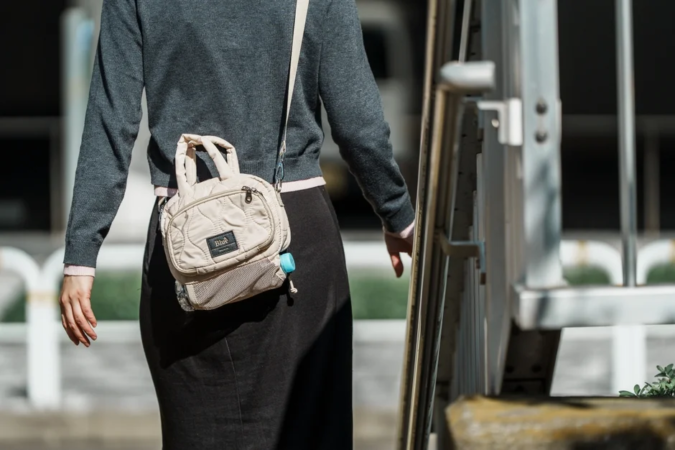 グレーのセーターと黒いスカートを着用した女性が、キルティングのベージュのショルダーバッグを肩から下げて屋外を歩いている後ろ姿です。バッグには小さなペットボトルが取り付けられています。
