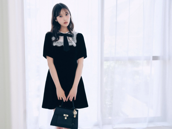 黒いレースの襟と黒いリボンが特徴の黒いワンピースを着た若い女性が、黒いハンドバッグを持って立っているポートレート写真です。