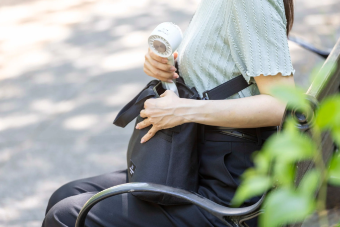公園のベンチに座った女性が、使用していた携帯扇風機を黒いカバンにしまっている瞬間
