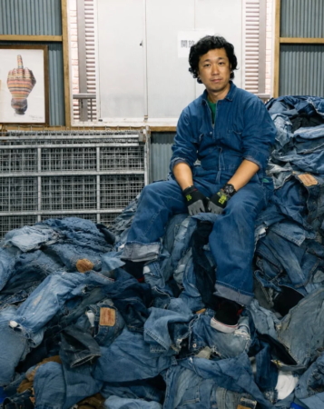 デニムのつなぎを着てジーンズの山の上に座る男性（RYOJI YAMASAWA）