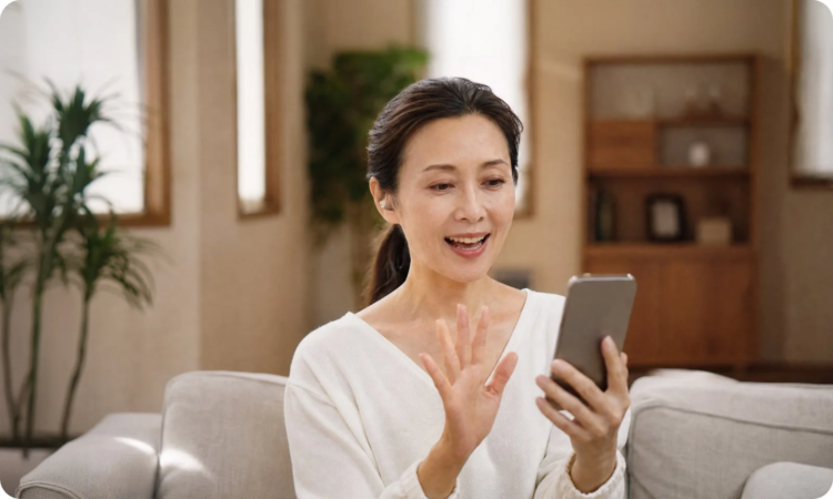 自宅のソファに座った女性が、スマートフォンを使って笑顔でビデオ通話を楽しんでいる様子