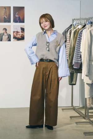 グレーのニットベストと水色のシャツ、茶色のワイドパンツを着用した女性