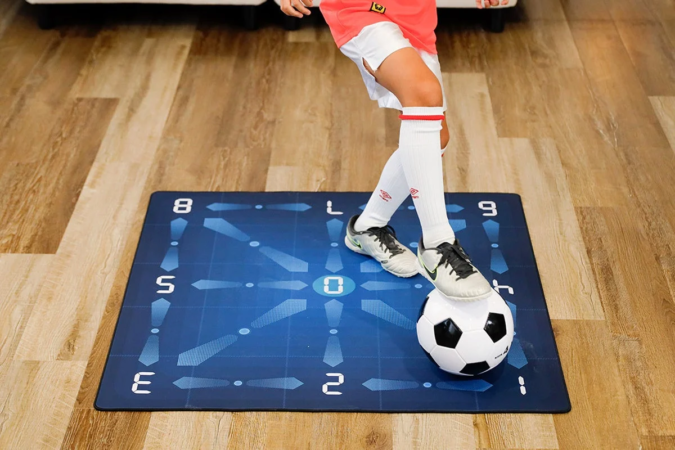 木製の床に広げられたサッカー練習用マットの上で、少年がサッカーボールに足を乗せている