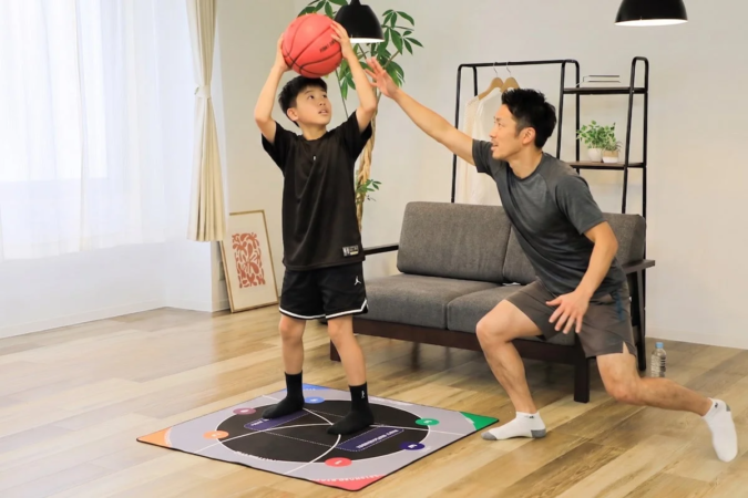 明るいリビングで、少年が赤いバスケットボールを持ち、大人が指導する様子