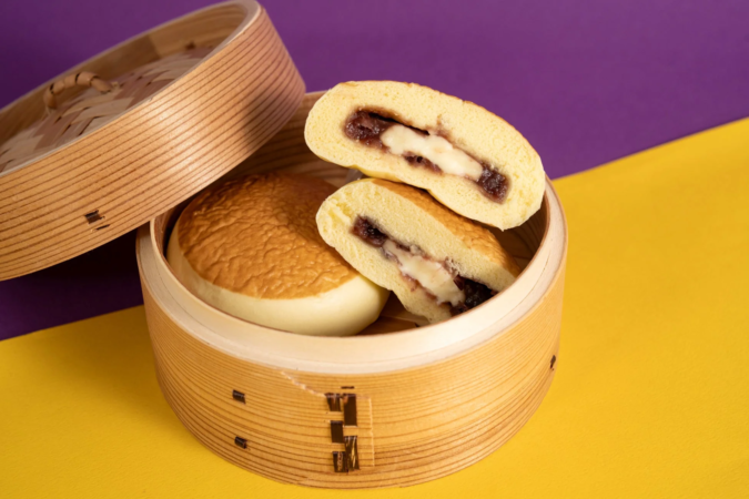 竹製の蒸し器に入った饅頭が写っています。手前の饅頭は半分に切られ、中には小豆餡とクリームチーズが詰まっているのが見えます。背景は紫と黄色の鮮やかなツートンカラーで、美味しそうな和スイーツの様子を伝えています。
