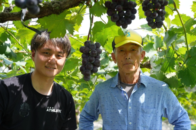ブドウ畑で、たわわに実ったブドウを背景に、若い男性と年配の男性の二人が並んで立っています