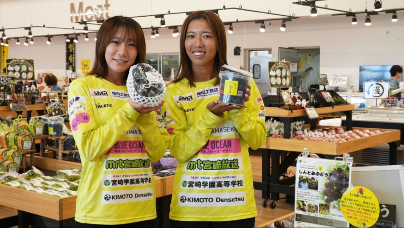 スーパーマーケットの青果売場で、黄色いユニフォームを着た2人の女性が、宮崎県産の「しんとみぶどう」を笑顔でPRしています